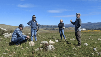 Four people in casual clothing stand in a semi circle in a green meadow with rocks talking to each other under a clear blue sky