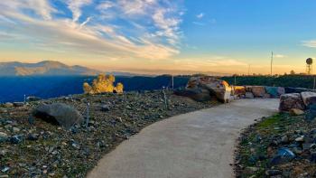 A paved trail curves around a rocky hilltop with a magnificent view of blue mountains all around, under a bright blue sky with a hint of yellow on the horizon