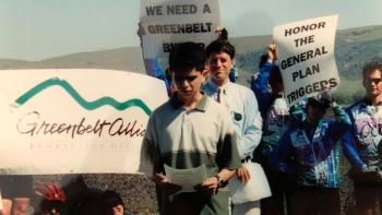 A teenager stands in front of a crowd holding signs about protecting Coyote Valley, looking down at a paper he is reading