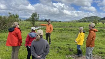 Six people in rain jackets stand in a loose circle in a green meadow, looking towards the left where the person in the center of the image is pointing