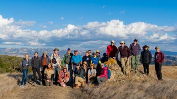 A group of 22 people in hiking clothes stand on the top of a brown grass-covered hill smiling at the camera, behind them is a blue sky filled with white fluffy clouds and golden mountains on the horizon