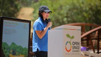 A woman in a black baseball hat, blue shirt, and sunglasses stands at a podium with the OSA logo speaking into a microphone at an outdoor event