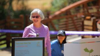 A woman in a pink shirt and sunglasses stands at a podium with the OSA logo speaking into a microphone at an outdoor event