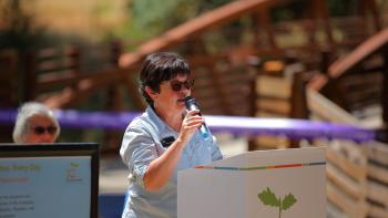 A woman in a light blue shirt and sunglasses stands at a podium with the OSA logo speaking into a microphone at an outdoor event