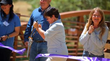 A woman in a light blue shirt cuts a purple ceremonial ribbon in front of a rusty red bridge, while people around her smile and clap