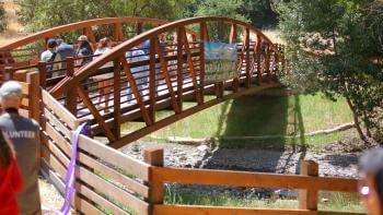 People walk across a rusty red pedestrian bridge over a low creek and trees