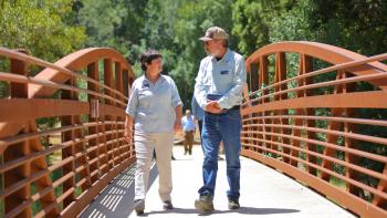 A pair of people wearing OSA branded clothing walk across a rusty red bridge towards the camera, talking to each other