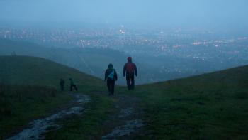 A pair of hikers wearing raincoats walks away from the camera down a wet and muddy trail, with city lights below them through a misty haze