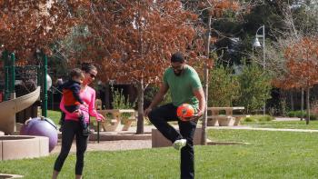 A woman wearing a pink shirt and sunglasses and holding a toddler on her hip smiles and points at the man next to them wearing glasses and a green shirt and juggling an orange soccer ball, at a small park lawn next to a play structure