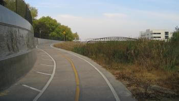 A paved bike trail with yellow and white lines curves up a slight incline with tall grasses on the right and a concrete wall with relief sculptures and a fence top on the left