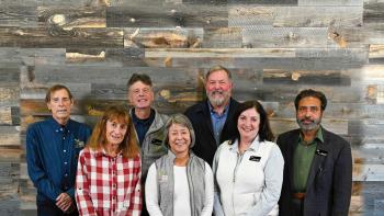 Group photo of seven people standing in front of a wood paneled wall, smiling at the camera