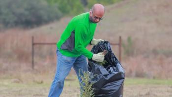 A land steward holds a trash bag while collecting invasive plants in a field