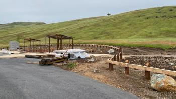 A central staging area is under construction with green hills in the background