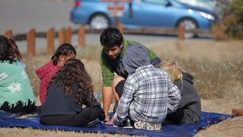 Students sit on a blanket while an instructor crouches down to teach them