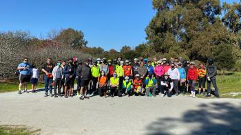A group of about 50 people wearing bicycle helmets standing on a gravel trail in front of trees and vegetation standing and kneeling while smiling at the camera