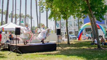 A pair of dancers wearing white traditional Mexican dresses with colorful ruffles perform on a small black stage in a park with tall green trees and festival booths in the background