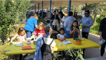 A ground of students sit around a yellow picnic table next to an outdoor school hallway, full of adults and other students. The students around the table are doing crafts, surrounded by trees and gardens