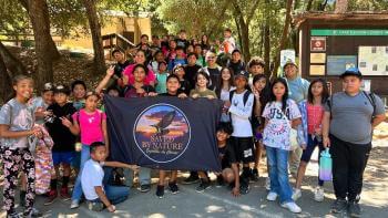 A group of about 30 middle school aged students stand in a natural park area facing the camera and holding up a large dark flag with a circular emblem with the Saved By Nature logo of an eagle and sunset