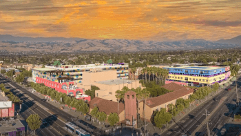 A drone shot of a city block corner with large, colorful buildings and a street lined with green trees, beyond in an orange sunset sky and golden hills in the distance