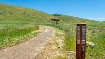 A wooden trail marker in the foreground in front of a compacted tan trail leading through a green field towards a covered picnic area, with green hills behind under a bright blue sky