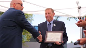A man in a dark blue suit holding a framed certificate with signatures reaches out to shake the hand of another man in a dark blue suit and glasses who has his back o the camera  