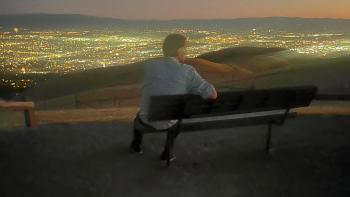 A man sits on a bench overlooking a vast cityscape below full of bright lights under a light purple twilight sky; his back is to the camera and his right arm is over the back of the bench, as he turns slightly to the right to take in the view 