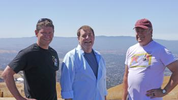Three men stand looking at the camera and smiling, in front of a view behind them with golden rolling hills, a city below, and blue mountains in the distance, under a clear blue sky