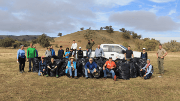 A group of 21 people stand and sit around a pile of large black trash bags filled with plant material in front of a brown grass-covered hill, under a blue and gray sky