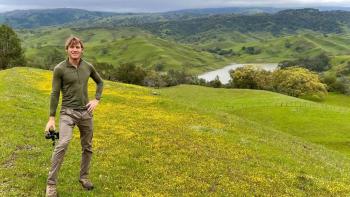 Man standing in a vast field of green and yellow grass with hills in the background