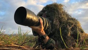 Close shot of a man laying on the ground, holding a camera, wearing a camouflage mossy ghillie suit  