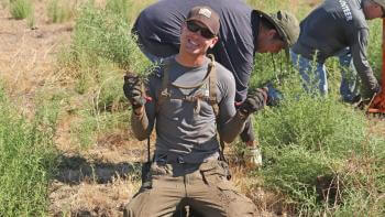 Close shot of man kneeling in dirt, holding weeds in both hands and smiling