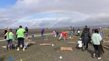 a group of children and adults walking around and helping to clean and restore the flat land. The sky is blue with grey clouds and a wide, full rainbow is in the background.