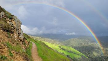 A dirt trail on the edge of a cliff with green grass on the right side, in front of a vast view of green mountains with a large rainbow spanning a dark gray and blue sky