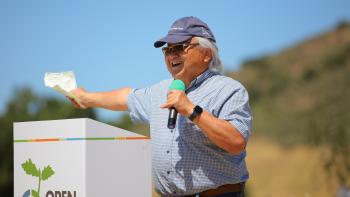 A person wearing a blue shirt and cap stands behind a white podium holding a black and green microphone and stretching an arm out, smiling
