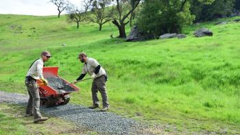 Wide view of two men shoveling rocks onto a trail surrounded by green hills