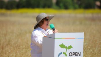 A person wearing a white shirt and broad rimmed hat stands behind a white podium holding a black and green microphone in front of a field of tall golden grass
