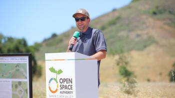 A person wearing a gray shirt and cap stands behind a white podium holding a black and green microphone, smiling