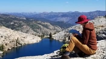 Wide shot of a woman sitting on a hill looking out at a blue lake and mountains