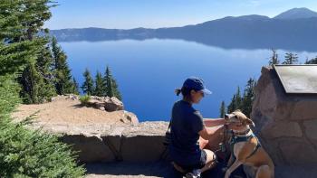 Wide image of woman kneeling on the floor with her dog in front of a blue lake and tall green trees 