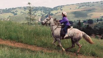 young girl riding a white horse on a trail surrounded by green grass and large hills