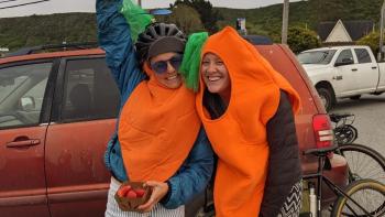 two women smiling in carrot costumes in front of a red car