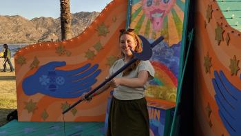 woman smiling and holding a large wand in front of a large and colorful art mural