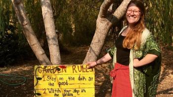 woman in front of a tree smiling while touching a yellow sign that reads "garden rules."