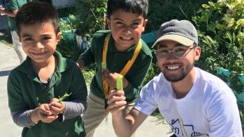 Two young students and a man smiling holding veggies from the garden