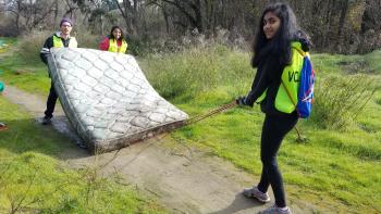 Three people pulling a soiled mattress through a dirt path surrounded by greenery.