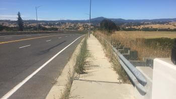 A street and sidewalk view with golden hills and a blue sky