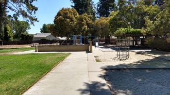 A park with a concrete sidewalk floor and a sand pit next to a play structure