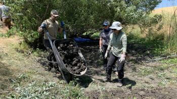A person dumps a wheelbarrow full of dirt into a dry streambed, standing next to two other people who are guiding the action