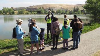 A group of people listening to a speaker in front of a lake