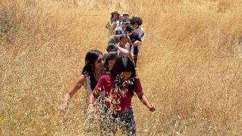 A group of students hikes through tall, dry grass in a scenic, open field up a slight hill towards the camera.
